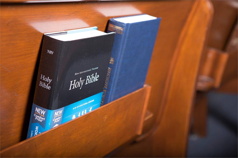 Two black NIV Pew and Worship Bibles standing in the book rack of a wooden church pew.