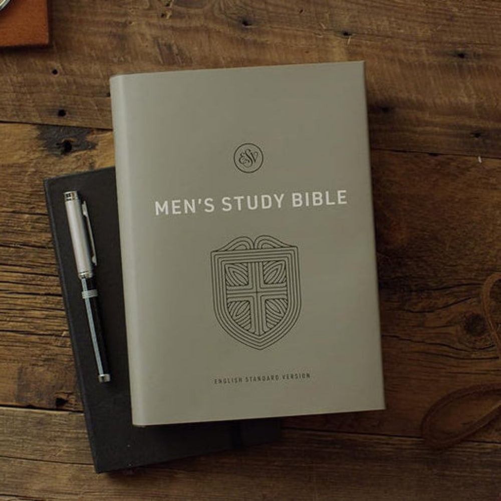 Gray-green ESV Men's Study Bible lying closed on a wooden desk with reading accessories.
