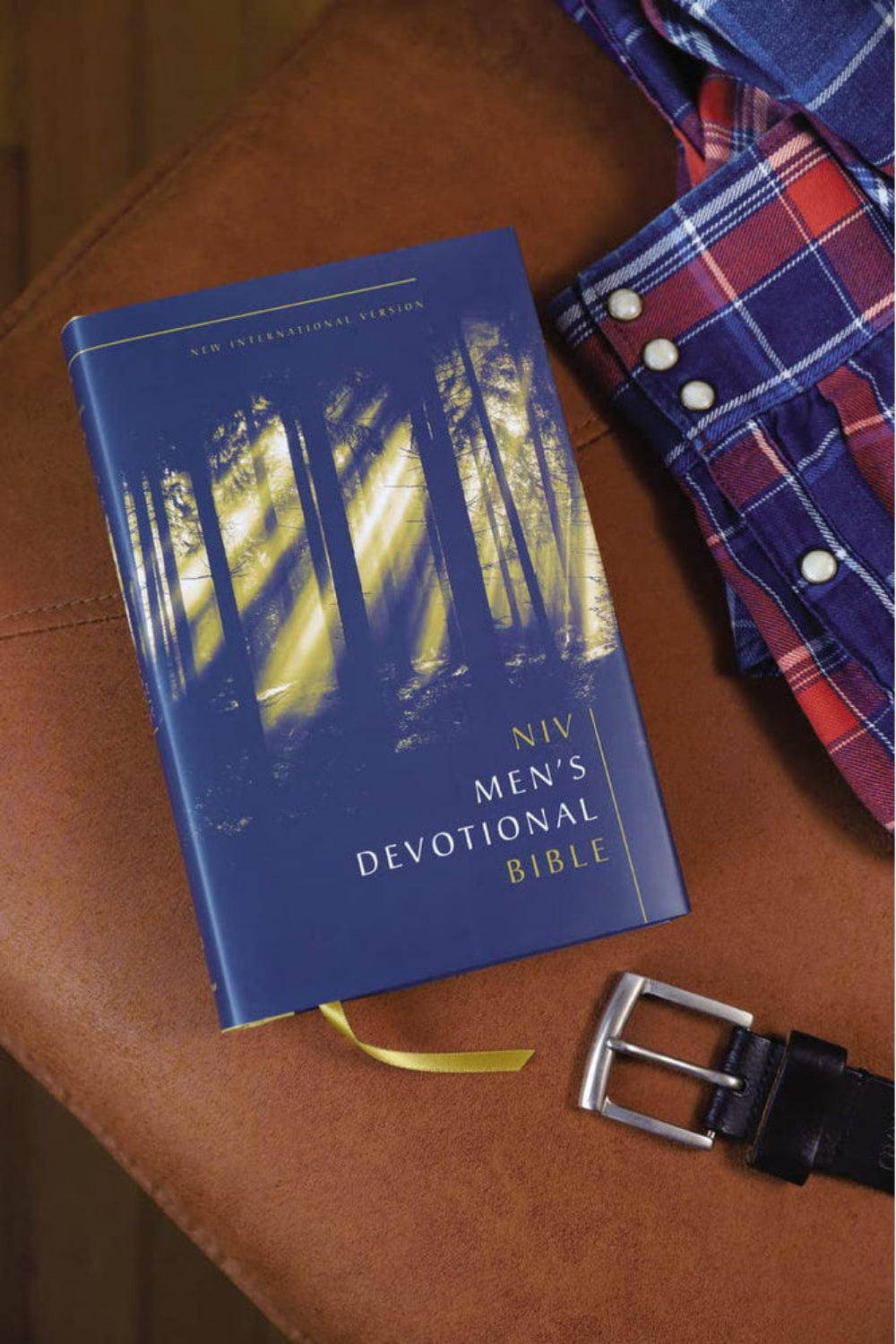 Navy NIV Men's Devotional Bible resting on a brown surface beside a plaid shirt and belt.