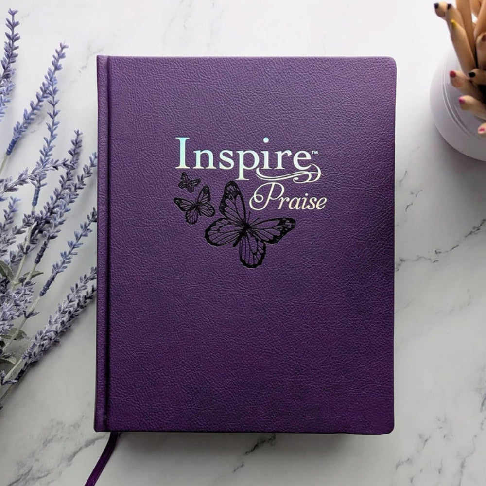Purple NLT Inspire Praise Bible resting closed on a pale tabletop beside lavender.